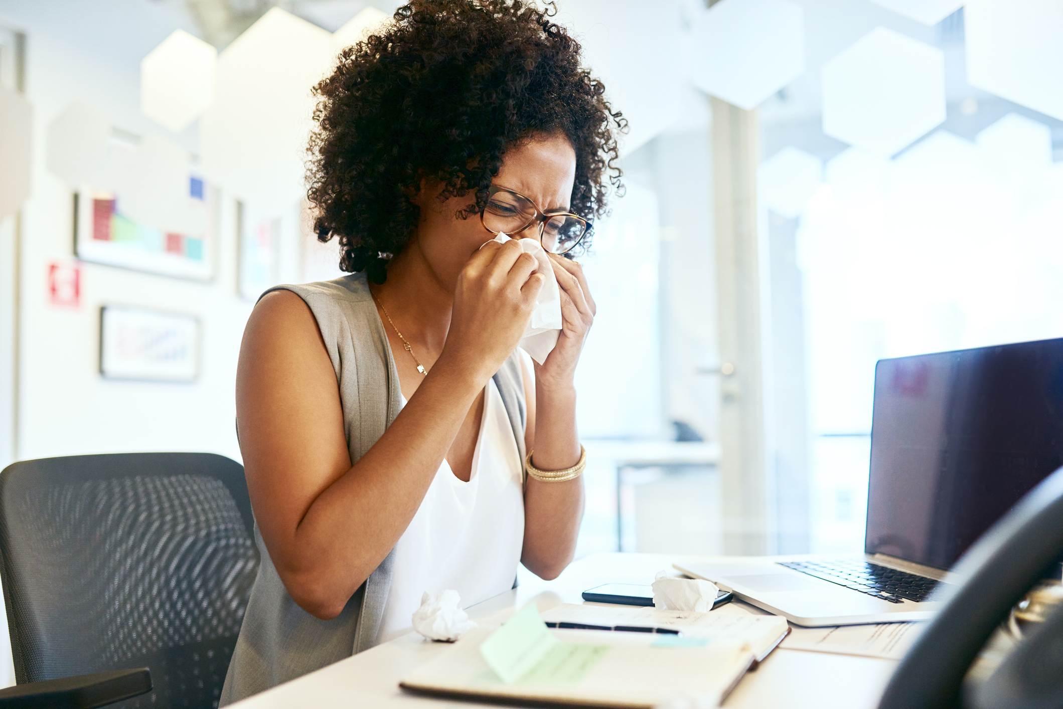 A sick woman blowing her nose at her desk in an office.